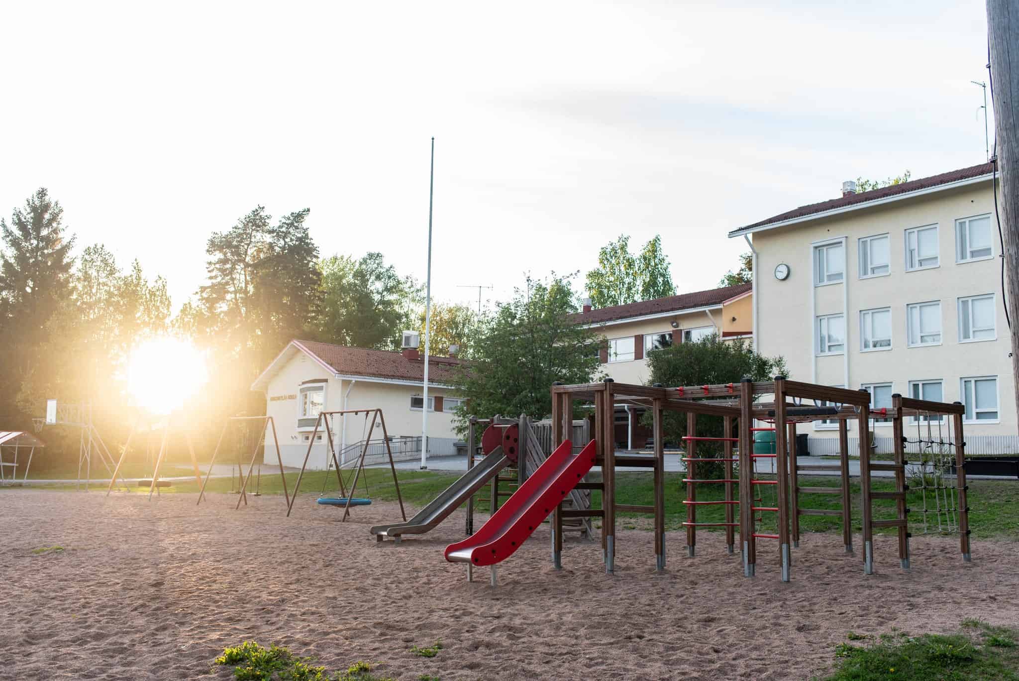 Rakennus, piha-alue, kiipeilyteline, keinut, kaksi liukumäkeä, hiekkaa, nurmikkoa.