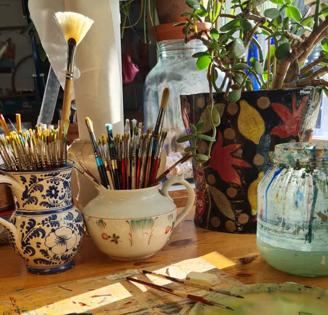 Työhuoneen pöydällä maalisutit purkeissa, taustalla keraaminen itsetehty kukkaruukku jossa keltaisia sitruunoita ja punaisia kukkasia.