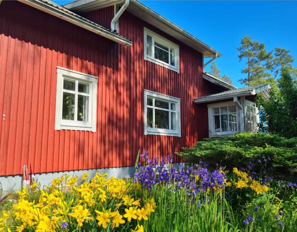 "Punainen puutalo puutarhassa sinisiä, keltaisia ja valkoisia kukkia."